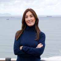 Una mujer con el pelo largo y castaño, vestida con un jersey de cuello alto azul oscuro y con los brazos cruzados.