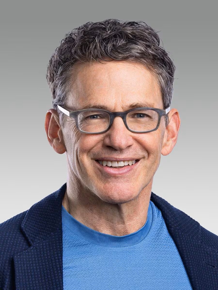 Portrait of a smiling man wearing glasses and a blue crew neck shirt with a suit jacket
