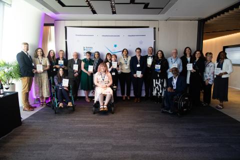 Winners of the 2025 awards smiling and holding their awards in front of a screen showing the Disability Smart Awards 2025 logo. Three people at the front are wheelchair users.
