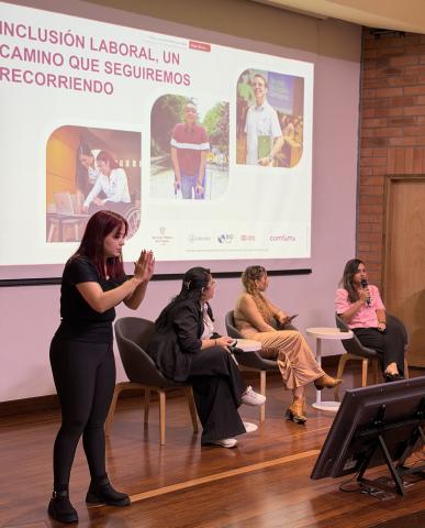 Tres personas sentadas en el escenario con una gran pantalla detrás de ellas y un intérprete de lengua de signos a un lado.