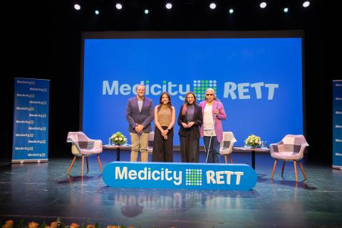 Cuatro personas de pie en un escenario con una gran pantalla azul al fondo y flores en el escenario. 