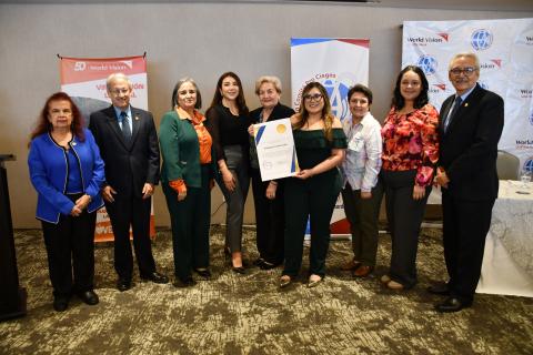 Nueve personas sonriendo mientras sostienen un certificado con tres carteles detrás de ellas. 