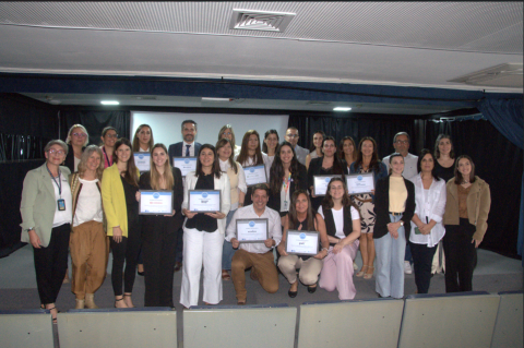 Varias personas sonriendo y sosteniendo certificados en un escenario con una gran pantalla blanca detrás de ellas. 