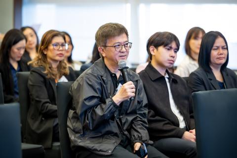 ○	A man sitting in rows of chairs in the audience holds a microphone as he asks a peer leader speaker questions about inclusion as a strategic framework for businesses.
